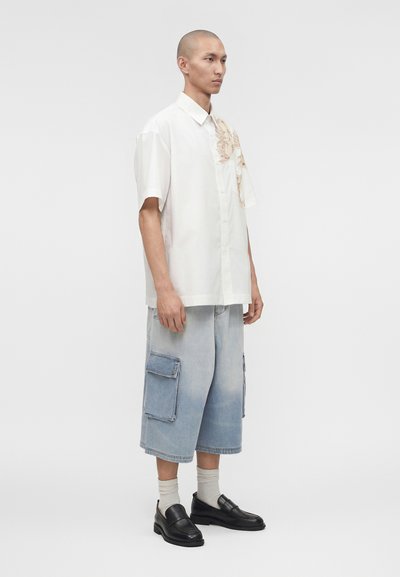 Άνδρας με ξυρισμένο κεφάλι που φοράει λευκό κοντομάνικο πουκάμισο με λουλουδάτη λεπτομέρεια, πολύ φαρδιά ανοιχτόχρωμα τζιν σορτς με τσέπες, λευκές κάλτσες και μαύρα μοκασίνια.