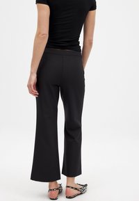 Črne široke hlače iz gladkega materiala z ravnim pasom. Zadnji pogled prikazuje prilegajoč fit in razširjen rob, v kombinaciji s črtastimi sandalmi.