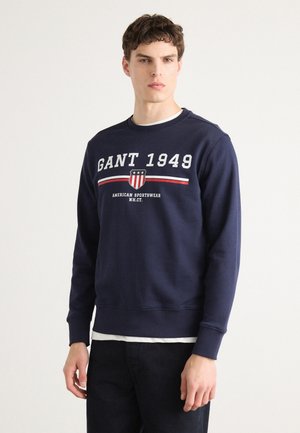 Navy blue cotton sweatshirt featuring "GANT 1949" text, a shield logo, and red accent stripes. Ribbed cuffs and hem, casual fit.