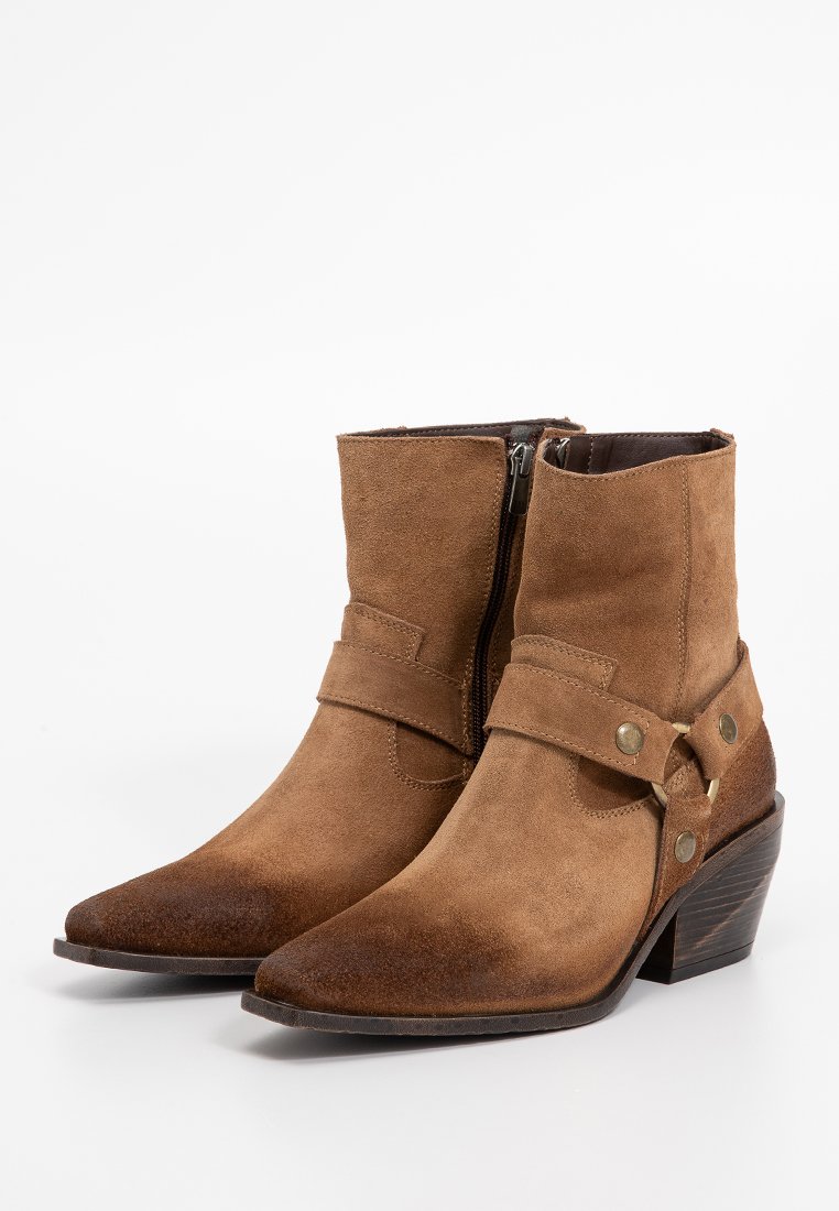 Botas de tobillo de ante marrón con punta afilada, cremallera lateral y detalles de correa con herrajes metálicos. Tacón bajo y apilado, parte superior texturizada.