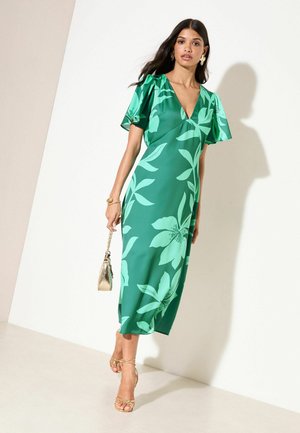 Kvinde iført en grøn midikjole med blomsterprint og flagrende ærmer, der holder en gylden taske og har gyldne remsandaler og øreringe på.