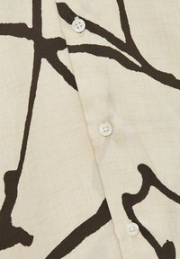Camisa de tela blanca con un audaz patrón abstracto en marrón, con un detalle de botón de plástico en la tapeta. Textura suave en general.