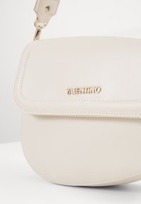 Beige läderhandväska med rundad form, magnetiskt lockdown och guld "Valentino"-logotyp. Slät yta och minimalistisk design.