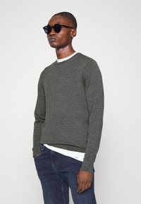 Pullover verde lavorato a maglia in lana, con scollatura rotonda e maniche lunghe, indossato sopra una camicia bianca e abbinato a jeans scuri.