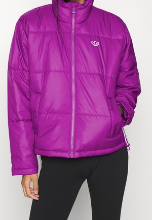 Fioletowa pikowana kurtka z przednim zamkiem i logo Adidas, noszona na czarnych spodniach, jedna ręka w kieszeni, druga opróżniona przy boku.