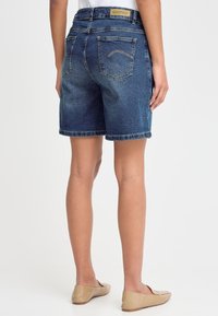 Femeie purtând pantaloni scurți de blugi albaștri de lungime medie și pantofi bej cu talpă joasă, stând cu spatele spre privitor pe un fundal alb.