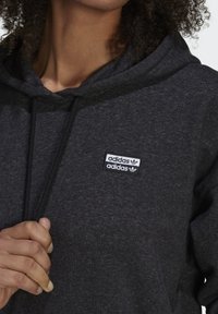 Czarna bluza z kapturem wykonana z miękkiego materiału z dużą przednią kieszenią. Na górnej lewej stronie klatki piersiowej znajduje się podwójna naszywka z logo Adidas.