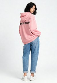 Felpa oversize rosa con testo nero "PARIS" sulla schiena, abbinata a jeans azzurri con risvolto e sneakers bianche. Tessuto di cotone.