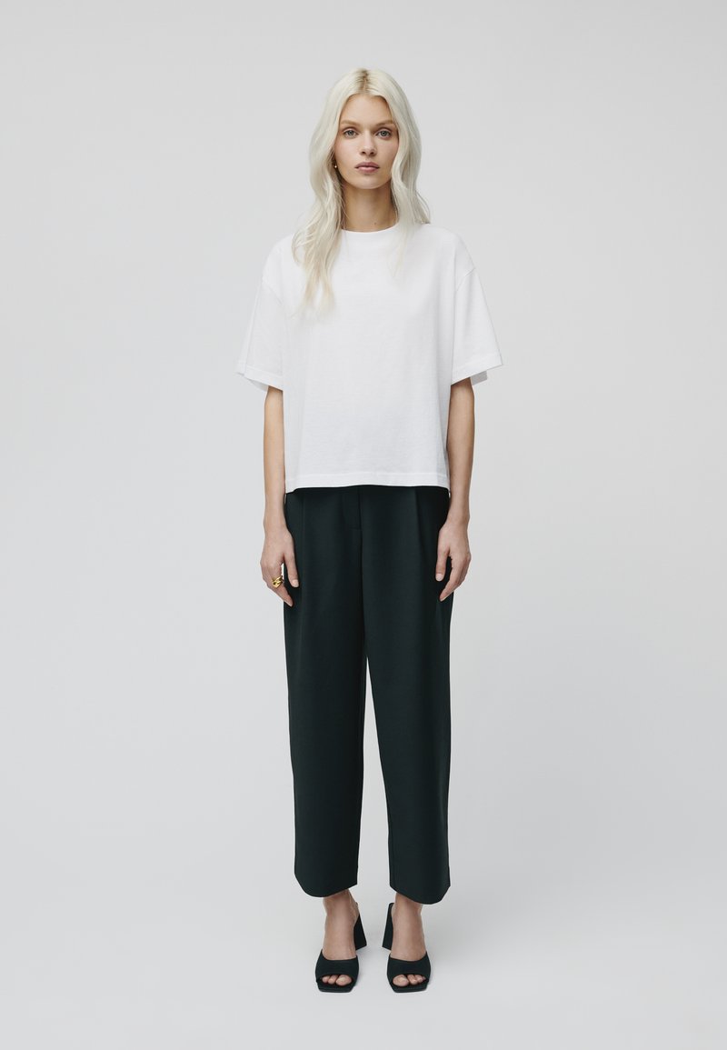 Weißes, kurzes T-Shirt mit kurzen Ärmeln, kombiniert mit lockeren, dunkelgrünen Hosen. Das Modell trägt schwarze, hochhackige Sandalen und einen auffälligen Ring.