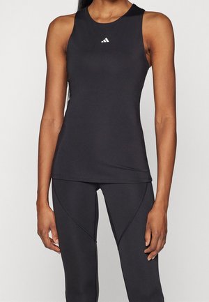 Vrouw draagt een nauwsluitend zwart mouwloos topje met een klein wit Adidas-logo op de borst en bijpassende zwarte legging met naaddetails.