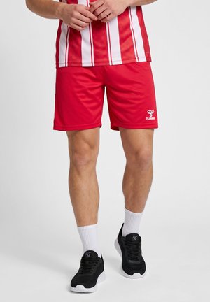 Man draagt rode sportshorts, een rood-wit gestreept shirt, witte sokken en zwarte sportschoenen, staand met zijn handen gevouwen.