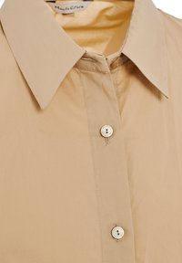 Camicia color beige con colletto a punta, dotata di chiusura a singolo bottone e realizzata in tessuto leggero e liscio. Dettagli di cucitura sottili visibili.