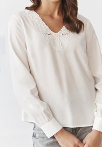 Blusa bianca con scollatura a V caratterizzata da ricami intricati e maniche lunghe. Il tessuto è liscio con un leggero lucore.
