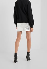 Donna che indossa una felpa nera, una mini gonna metallica argento e stivaletti neri con tacco, in piedi su uno sfondo bianco semplice.