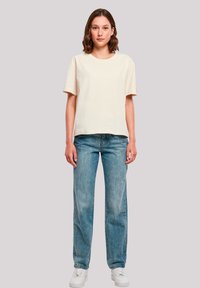 Helles beige Baumwoll-T-Shirt mit kurzen Ärmeln und Rundhalsausschnitt; kombiniert mit geraden, hellblauen Denim-Jeans und weißen Sneakers.