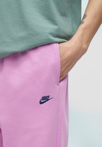 Osoba s rukou vo vrecku fialových teplákov Nike, oblečená v zelenej košeli, ukazujúca tetovanie na predlaktí na svetlom pozadí.