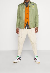 Lys grøn jakke, orange cardigan, ternet skjorte, beige cargo-bukser og hvide sneakers med sorte accenter og farverige detaljer.