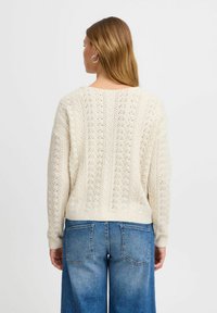 Krämfärgad stickad tröja med ett mönster och ribbad nederkant, kombinerad med blå vida jeans, sedd bakifrån.