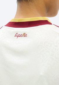 Άτομο που φορεί λευκό πουκάμισο με κόκκινο και κίτρινο γιακά, με κεντημένο το "España" σε κόκκινο και κίτρινο στο πάνω μέρος της πλάτης.