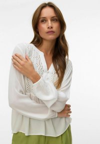 Femme aux longs cheveux bruns portant un chemisier blanc à broderies ajourées et un pantalon vert, les bras doucement croisés sur sa poitrine.