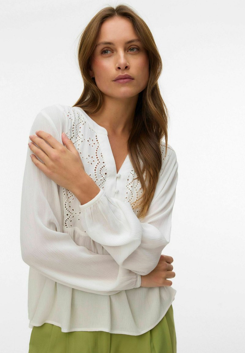 Femme aux longs cheveux bruns portant un chemisier blanc à broderies ajourées et un pantalon vert, les bras doucement croisés sur sa poitrine.