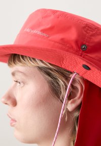 Chapeau seau rouge en tissu léger. Présente un large bord, une sangle de menton ajustable avec un cordon rose et des détails de logo sur le devant.