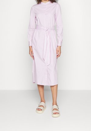 Žena v růžově-bílých pruhovaných midi šatech s dlouhým rukávem, se zavázaným páskem v pase a bílými sandály na platformě stojí před jednobarevným pozadím.