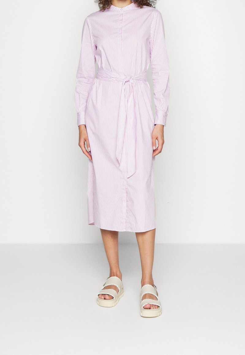 Femme portant une robe midi à manches longues rayée rose et blanche avec une ceinture nouée à la taille et des sandales à plateforme blanches, debout devant un fond uni.