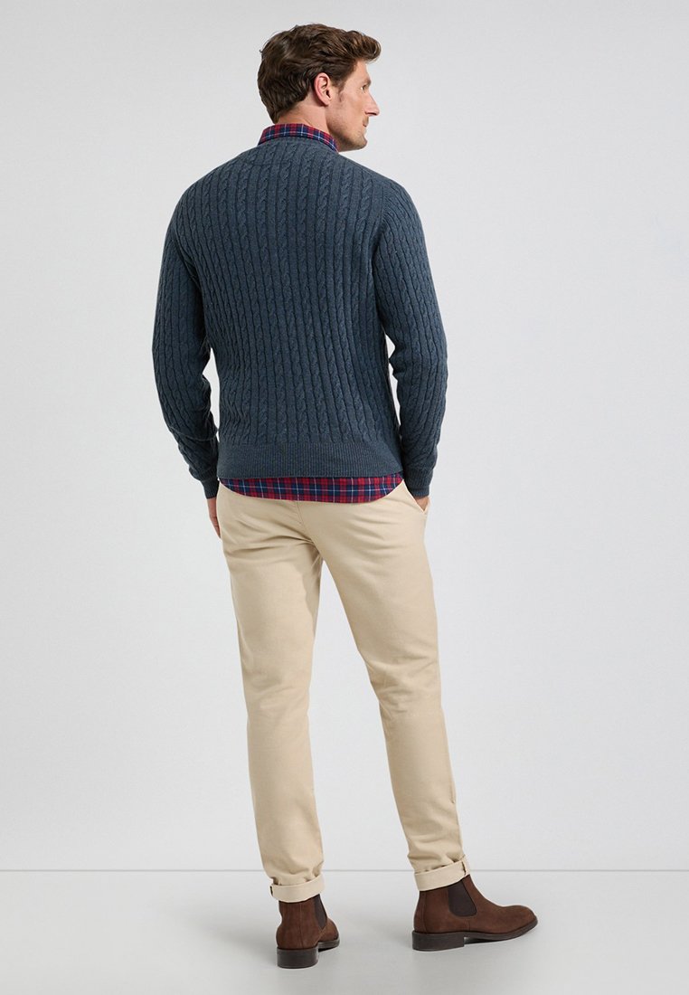 Donkerblauwe kabeltrui over een geruite shirt, gecombineerd met beige broek en bruine enkellaarsjes. Heeft een gestructureerde brei en een casual ontwerp.