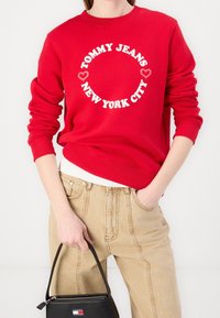 Persona vistiendo un sudadera roja con texto circular en blanco y rosa "Tommy Jeans New York City" y pantalones beige, sosteniendo un pequeño bolso negro.