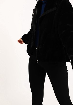 Persona que lleva una chaqueta de piel sintética negra, un suéter de cuello alto azul marino y pantalones negros, mostrada desde los hombros hasta la parte superior de los muslos.