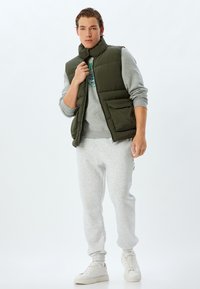 Oliven grøn polstret vest over en grå sweatshirt, parret med grå joggers og hvide sneakers; har en høj krave og frontlommer.