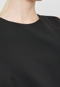 Nahaufnahme eines weiblichen Halses und Oberkörpers, der ein schlichtes schwarzes ärmelloses Top vor einem neutralen Hintergrund trägt.