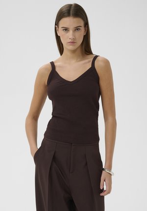 Noor naine, kes kannab tumedat pruunikat soonikutanktoppi ja sobivaid laia säärega pükse, seisab käed taskutes tavalise tausta ees.