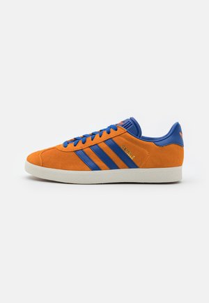 Pomarańczowy zamszowy sneaker z niebieskimi paskami, niebieskimi sznurowadłami i białą podeszwą, widok z boku na białym tle.