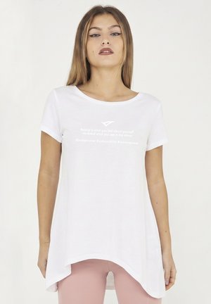 Jonge vrouw met steil haar die een wit asymmetrisch T-shirt met inspirerende tekst en roze broek draagt, staand tegen een effen achtergrond.