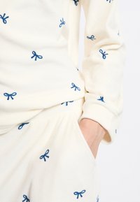 Conjunto de pijama de forro polar crema con bordado de lazo azul; presenta mangas largas, un corte relajado y bolsillos en los pantalones. Textura suave.