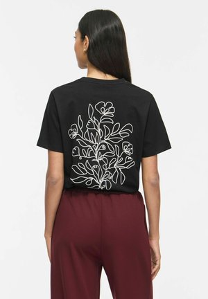 Kvinde med langt mørkt hår iført en sort t-shirt med hvidt blomsterlinjekunst på ryggen og mørkerøde bukser, stående med ryggen til.