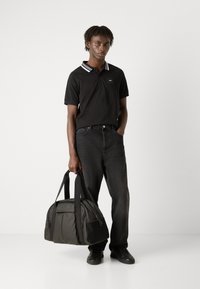 Camisa polo negra con cuello a rayas blancas, jeans negros y una bolsa de viaje oscura con una marca prominente. Un atuendo casual con elementos de diseño sencillos.
