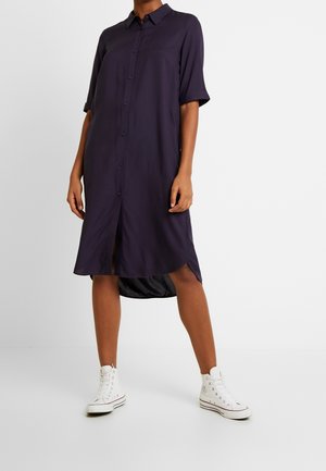 Osoba ubrana w ciemnofioletową koszulową sukienkę z podwiniętymi rękawami oraz białe trampki wysokie, stojąca na białym tle.