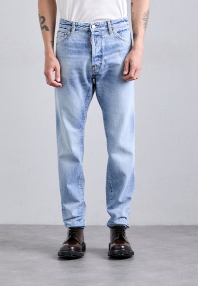 Person iført lyseblå slidte jeans og mørkebrune snørebåndsstøvler, som står på et gråt gulv mod en ensfarvet lys væg.