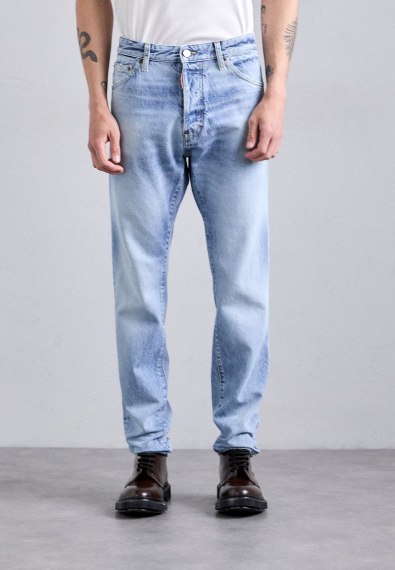 Persona con jeans descoloridos de color azul claro y botas de cuero marrón oscuro con cordones, de pie sobre un suelo gris frente a una pared clara y sencilla.