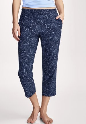 Woman wearing navy blue cropped pants with light blue dotted swirl pattern, hands in pockets, standing barefoot on a light background.