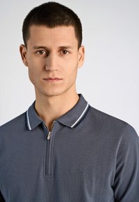 Graues Poloshirt mit einem Reißverschluss am Kragen, weißen Streifenakzenten und dezent strukturiertem Stoff. Mit einem aufgestickten Logo auf der Brust.