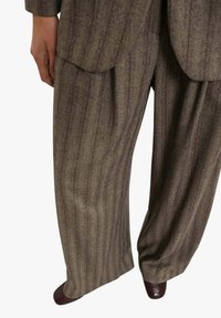 Pantaloni di lana a righe grigie e marroni con una vestibilità ampia e pieghe. Il tessuto ha una texture morbida; abbinati a scarpe marrone scuro.