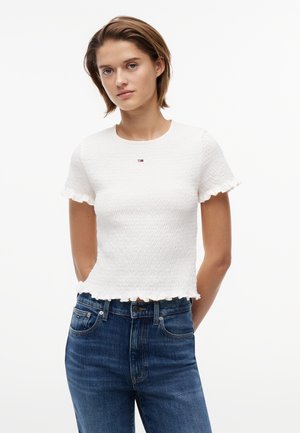 Νεαρή γυναίκα με κοντά μαλλιά, που φοράει ένα λευκό υφασμάτινο μπλουζάκι με κοντά μανίκια και μπλε τζιν, στέκεται με τα χέρια πίσω από την πλάτη.