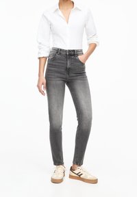 Mujer con camisa blanca de botones y mangas arremangadas, jeans skinny de tiro alto en un negro deslavado y zapatillas beige con toques marrones.