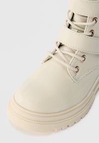 Skórzany botek w kolorze kremowym z płaskimi sznurowadłami, metalowymi oczkami i grubą, teksturowaną podeszwą. Posiada poziome paski na górze.