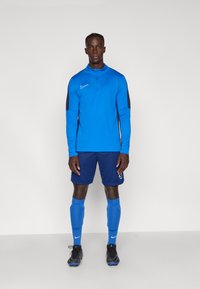 Μπλε μπλούζα με μακριά μανίκια και φερμουάρ, μαύρες λεπτομέρειες; μπλε σορτς; ταιριαστά μπλε κάλτσες; μαύρες ποδοσφαιρικές μπότες με μπλε λεπτομέρειες; το λογότυπο της Nike είναι ορατό.