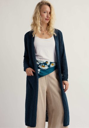 Blondýna v dlouhém tmavě modrém kardiganu, bílém tílku, béžových kalhotách a tyrkysovém šátku s bílými kopretinami uvázaném kolem pasu.